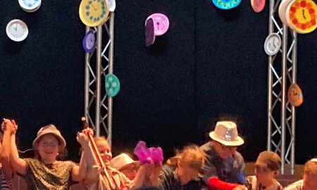 Von oben hängen viele bunte Uhren herab. Darunter verbeugen sich die Kinder. Sie stehen in einer Reihe und halten sich an den Händen.