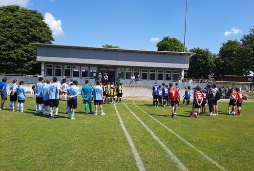 Circa 5 Schüler-Fußballmannschaften stehen mit dem Rücken zur Kamera auf dem Rasen und schauen Richtung Tribüne, wo zwei Personen stehen