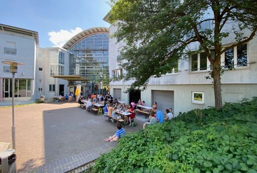viele Menschen sitzen vor einem Gebäude im Schatten an Biergarnituren und essen und trinken.