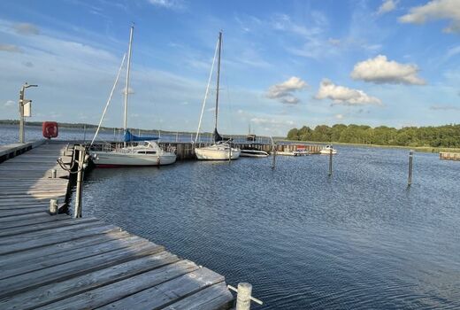 Ein Steg ragt ins Wasser. Er knickt nach rechts ab. An ihm haben zwei Segelboote festgemacht.