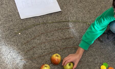 Auf dem Teppichboden liegen ein Zettel mit den Notenwerten C D E F G A H C, vier Äpfel, sechs dünne Äste und fünf kleine bunte Holzkreise.