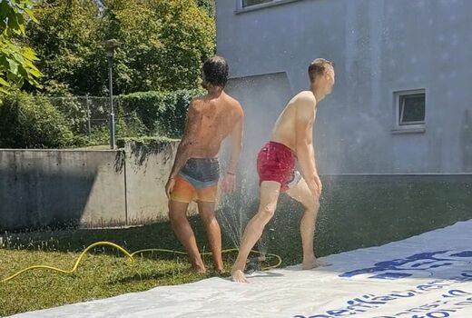 Auf dem Rasen liegt eine LKW-Plane. Links läuft ein Wassersprinkler. Zwei Personen stehen daneben.