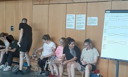 Schülerinnen und Schüler sitzen auf der Bank an der Wand. Sie halten ihre Badminton Schläger in der Hand. Rechts steht eine Flipchart auf der die Begegnungen zu sehen sind.
