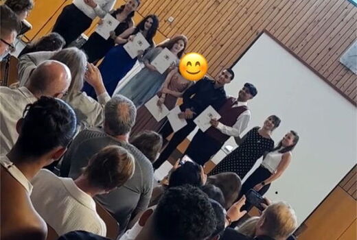 Schrägaufnahme: Im Vordergrund sitzen die Besucher der Feier und hinten lächeln die SchülerInnen der Klasse 9c in die Kamera.