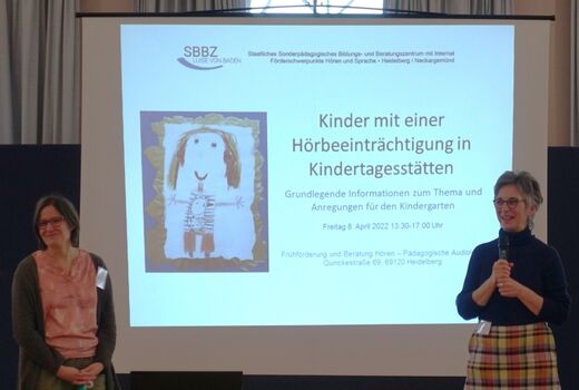 Zwei Frauen stehen mit Kikrofon vor einer Leinwand auf der eine Präsentation mit dem Titel "Kinder mit einer Hörbeeinträchtigung in Kindertagesstätten