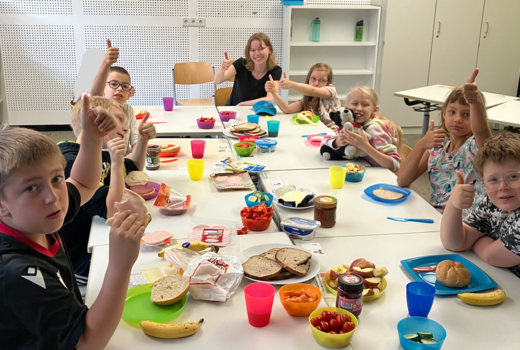 Eine Lehrerin sitzt mit sieben Kindern der zweiten Klasse im Klassenzimmer an einem langen und reich gedeckten Frühstückstisch. Alle zeigen einen Daumen hoch.