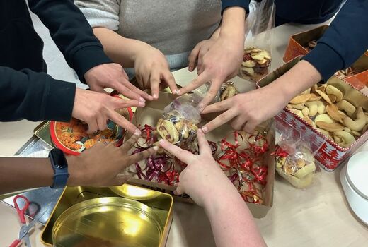 Auf einem Tisch werden Plätzchen eingetütet. Man sieht Dosen, Plätzchen, Geschenkband und sieben Schülerhände, die darüber mit gespreizten Zeige- und Mittelfinger einen Stern bilden