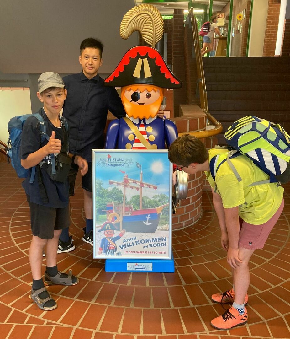Drei Schüler stehen neben einer großen Playmobil-Figur.