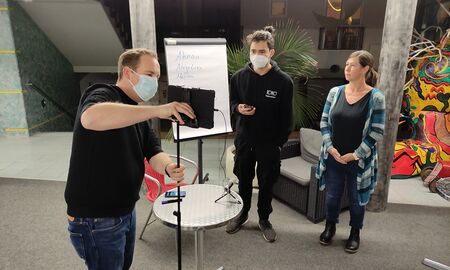 Herr Ulrich richtet die Kamera aus. Diese steht auf einem Stativ. Ein Schüler und Frau Edelmann stehen gegenüber.