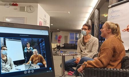 Eine Schülerin stellt sich vor. Herr Eisenbeiß sitzt neben ihr. Auf der linken Bildhälfte ist ein aufgeklappter Laptop zu sehen. Er zeigt die Übertragung des Live-Streams.