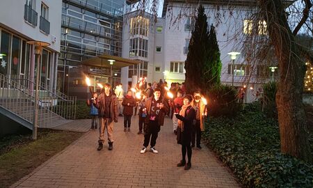 Alle haben sich vor dem Internatseingang versammelt, um zur gemeinsamen Wanderung aufzubrechen. Fast alle SchülerInnen und ErzieherInnen halten eine Fackel in der Hand. Im Hintergrund ist das Internatsgebäude zu sehen.