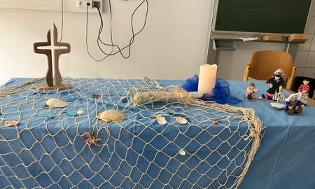 Ein Tisch mit einem blauen Tuch, einem Netz mit Muscheln und Seesternen, einer Flaschenpost, einer Kerze, einem Kreuz und Playmobil-Figuren ist als Altar geschmückt.