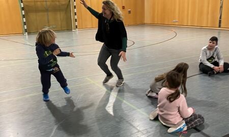 Ein Kind und eine Lehrerin tanzen in der Turnhalle. Drei weitere Kinder sitzen auf dem Boden