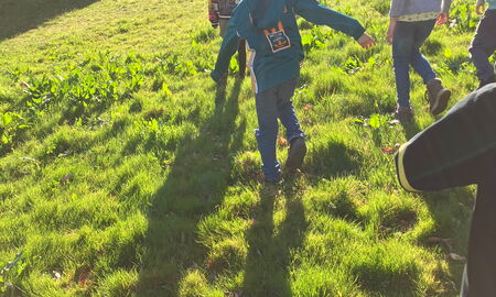Mittig im Bild sind drei Kinder draußen auf einer Wiese. Oben links sieht man ein Gebäude. Oben rechts sieht man einen Bauwagen.