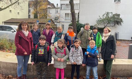 Zehn Schülerinnen und Schüler einer ersten Klasse posieren mit ihrer Lehrerin und ihrer Bezugserzieherin vor einem Baum in der Stadt für ein Gruppenfoto.