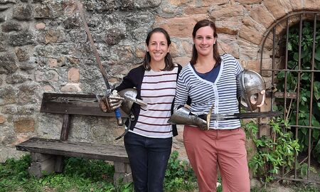 Zwei Klassenlehrerinnen posieren nebeneinander mit verschiedenen Ritterrequisiten wie Helm, Schwert und Rüstungshand vor einer alten Mauer.