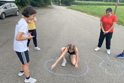 Vier Jugendliche befinden sich auf einer Straße. Ein Mädchen kniet und malt mit Kreide Figuren auf den Asphalt.