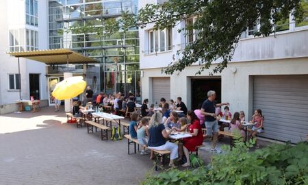 viele Menschen sitzen vor einem Gebäude im Schatten an Biergarnituren und essen und trinken.