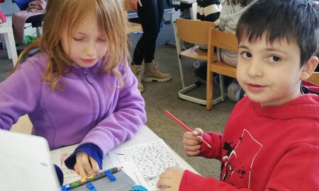 Ein Mädchen und ein Junge sitzen nebeneinander. Sie suchen auf einem Ausmalbild alle Felder mit dem Buchstaben M und malen diese farbig an. Im Hintergrund sitzen weitere Kinder.