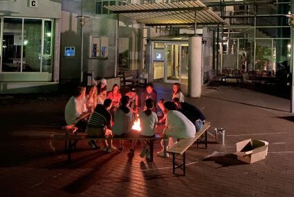 Eine Gruppe Jugendlicher sitzt am Lagerfeuer vor dem Internatsgebäude