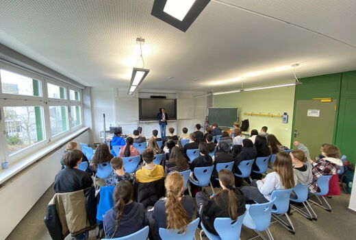 In einem großen Raum sitzen viele Schüler. Im Hintergrund steht Herr Katzenstein und stellt sich den Fragen der Schüler.