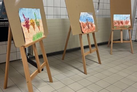 Im gefliesten Kunstraum stehen drei Staffeleien nebeneinander. Auf jeder Staffelei ist ein Bild ausgestellt.