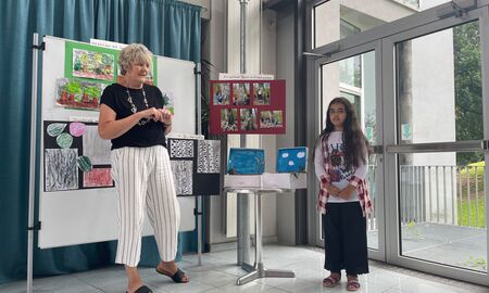 Eine Erzieherin und eine Schülerin stehen vorne vor verschiedenen Waldplakaten  und stellen einen Wald im Schuhkarton vor.