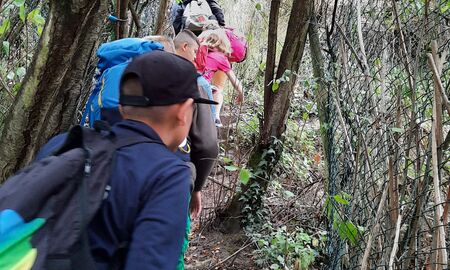 Kinder laufen hintereinander einen steilen und engen Waldweg hinauf. Rechts und links sind ein Maschendrahtzaun und Bäume.