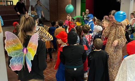 Auf der Bühne stehen Lehramtsanwärterinnen und tanzen zu Musik vor. Davor stehen und laufen verkleidete Kinder und deren auch verkleideten Lehrkräfte und tanzen und feiern.