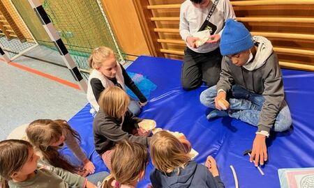 Acht Kinder sitzen gemeinsam auf einer Weichbodenmatte in einer Turnhalle