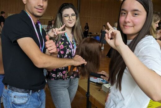 Ein Junge und zwei Mädchen stehen um einen Frisierkopf. Sie lächeln in die Kamera.