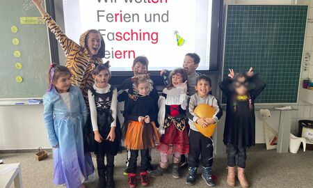 Eine als Tiger verkleidete Lehrerin posiert mit acht verkleideten Schülerinnen und Schülern im Klassenzimmer vor der Tafel.