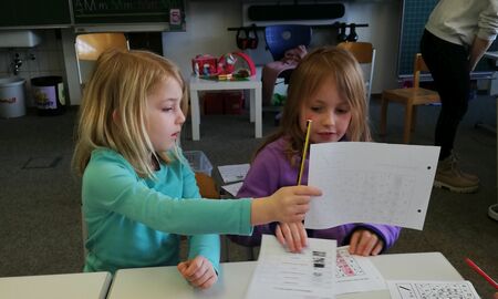 Ein Mädchen und eine Erstklässlerin sitzen nebeneinander. Hinter ihnen sieht man die Tafel. Das Mädchen zeigt der Erstklässlerin ihr Arbeitsblatt.