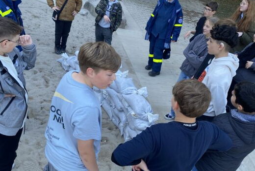 Schüler und zwei Helfer vom THW stehen um eine kleine, niedrige Mauer aus Sandsäcken.