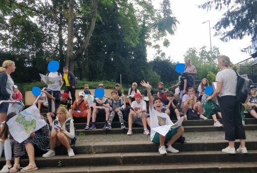 Die Kinder der fünften Klassen sitzen oder stehen auf den Stufen. Manche betrachten Faltpläne des Parks.