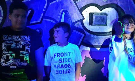 Drei SchülerInnen stehen nebeneinander. Der Junge links trägt ein schwarzes T-Shirt. Der Junge in der Mitte trägt ein weißes T-Shirt und lächelt. Ein Mädchen steht rechts. Sie hat den rechten Arm gehoben und lächelt.