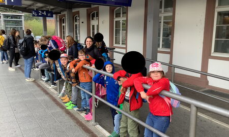 Zu sehen sind mehrere Kinder, die hinter einer Absperrung durch eine Stange nebeneinander an einem Bahnhof stehen und in die Kamera lächeln.