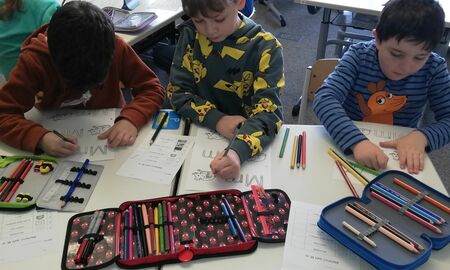 Drei Jungen sitzen gemeinsam an Tischen und schreiben mit Buntstiften den Groß- und Kleinbuchstaben M bunt. Im Hintergrund sitzen weitere Kinder an ihren Tischen.