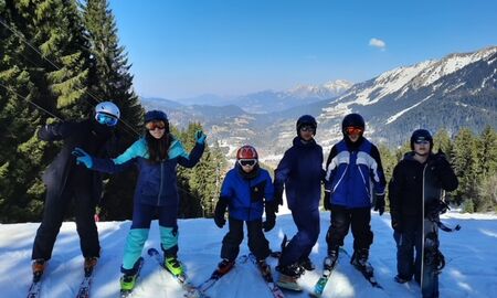 Sechs Schüler und Schülerinnen stehen auf Skiern und Snowboard im Schnee, im Hintergrund Berge und eine Ortschaft, die Sonne scheint