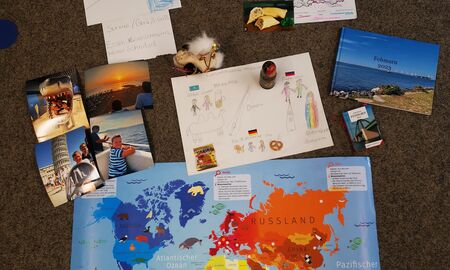 Unten liegt eine Weltkarte. Darüber liegen Urlaubsmitbringsel und Urlaubserinnerungen wie Fotos oder Fotobücher. Es gibt Informationen über unterschiedliche Länder oder Bundesländer und ein Plakat über verschiedene Herkunftsländer innerhalb einer Familie.