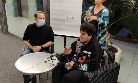 Schüler stellt sich sich vor. Dabei blickt er in die Kamera und spricht in ein Mikro. Neben ihm sitzt Herr Ulrich. Frau Edelmann steht im Hintergrung und dolmetscht.