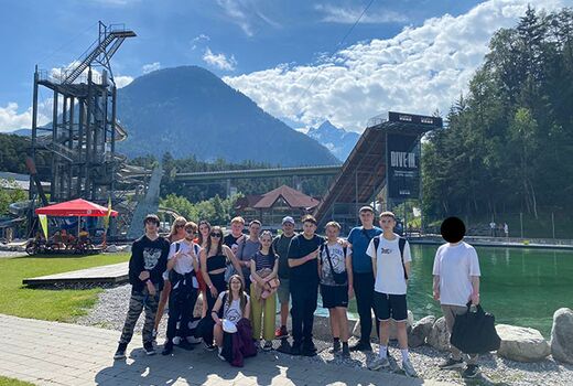 Ein Gruppenfoto vor dem großen Sprungturm der Area 47 mit einem Bergpanorama im Hintergrund.