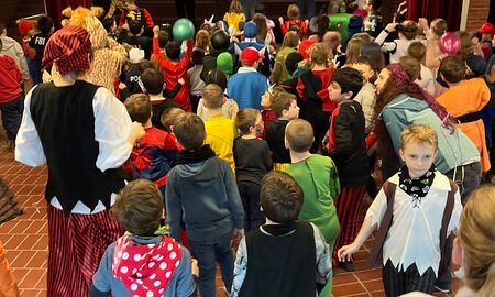 Auf der Bühne der Aula tanzen Lehramtsanwärterinnen vor und davor stehen die verkleideten Schülerinnen und Schüler der Grundschule Sprache.