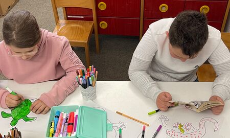Zwei Kinder sitzen nebeneinander an einem Tisch. Das Mädchen links malt eine Schildkröte grün aus. Der Junge rechts liest gerade ein Comicbuch.