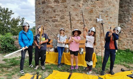 Eine Lehrerin und fünf Kinder posieren mit Ritterhelmen auf dem Kopf und Schwertern in den Händen vor einer Burgmauer.