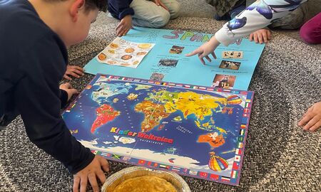 Fünf Erstklässler und Erstklässlerinnen sitzen auf dem Teppichboden um ein Plakat über Spanien und eine Weltkarte herum. Vorne stehen Tapas in einer Aluschale. Ein Mädchen zeigt auf das Plakat.