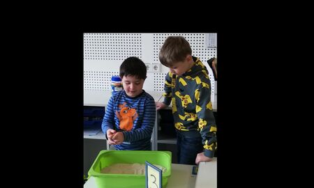 Ein Kind schreibt das M an einer Station in Sand in einer Kiste und ein anderes Kind hilft ihm dabei.