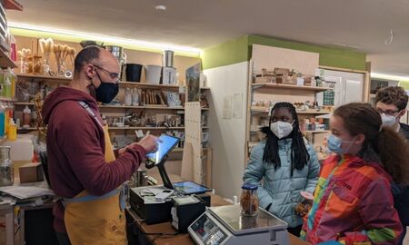 Ein Mann tippt den Preis für ein Schraubglas mit Lebensmitteln in ein Display, drei Kinder schauen zu