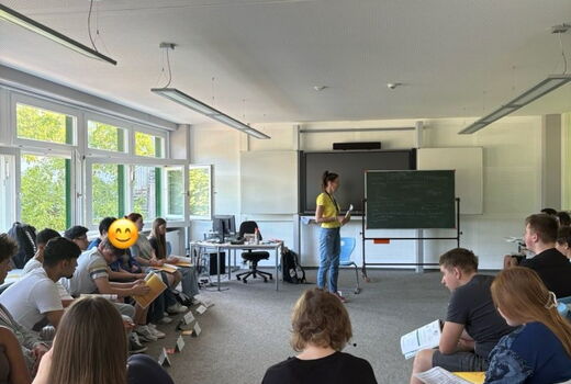 Blickin einen Raum, wo viele Schüler entlang der Wände sitzen. Sie hören einer Vortragenden zu.