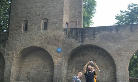 Der Lehrer fotografiert, zwei Schülerinnen schauen ihm aufmerksam dabei zu. Im Hintergrund die alte Stadtmauer mit Turm.